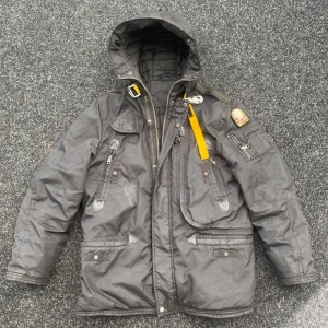 Parajumper - Grym svart parkajacka från Parajumpers med ikonisk gul rem vid kragen, patch på ärmen och flera fickor med dragkedjor och knappar. Jackan har huva, quiltat foder och coola detaljer i svart. Perfekt för kalla dagar när du vill se både chill och stilsäker ut.