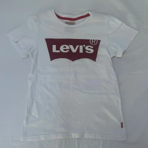 Vit Levi's t-shirt med röd logga - Vit t-shirt från Levi's med klassisk röd logga framtill. T-shirten har rund halsringning med röd detalj på insidan och korta ärmar. Tillverkad i mjuk bomull, perfekt för en avslappnad och stilren look.