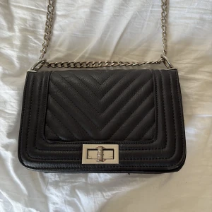 Svart quiltad med kedja - Snygg svart väska som går att ha både som axelväska och vanlig väska i skinnimitation med quiltad design och silvrig kedjerem. Väskan har ett stilrent spänne framtill och ett rymligt fack med innerficka och dragkedjefack. Perfekt storlek för det viktigaste och går att använda till allt!