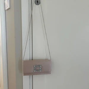Beige kuvertväska med strassdetalj - Elegant beige/rosa kuvertväska i satin med silverfärgad kedja och glittrande strassdetalj framtill. Perfekt storlek för det viktigaste och ger en lyxig känsla till din outfit. Enkel men ändå riktigt snygg design.