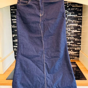 Mörkblå lång jeanskjol med fickor - Säljer en snygg mörkblå lång jeanskjol med klassiska kontrastsömmar i gult. Kjolen har fram- och bakfickor, bälteshällor och stängs med knapp och dragkedja framtill. Perfekt för dig som gillar en tidlös denimlook.