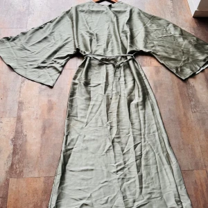 Lång grön kimono-klänning med bälte - Snygg lång kimono-inspirerad klänning i ljusgrön ton med breda ärmar och tillhörande knytbälte i midjan. Materialet har en mjuk, silkeslen känsla och faller snyggt längs kroppen. Perfekt för dig som gillar chill och flowy vibes.