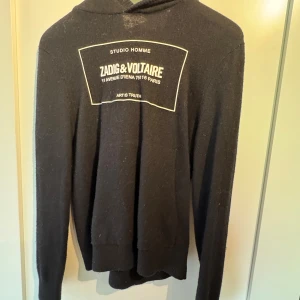 Svart hoodie från Zadig & Voltaire - Svart hoodie från Zadig & Voltaire med tryck på bröstet och mjukt stickat material. Tröjan har huva, långa ärmar och ribbade muddar. Den är S men sitter som xs