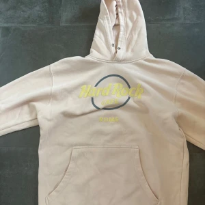 Beige Hard Rock Cafe hoodie Rome - Säljer en beige hoodie från Hard Rock Cafe med tryck i gult och grått på bröstet där det står 'Rome'. Tröjan har huva, känguruficka och är långärmad. Perfekt för dig som gillar streetwear och vill ha något unikt från en ikonisk plats.