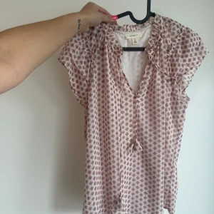 Blommig blus från Tamara Hill - Söt blommig blus från Tamara Hill i ljusrosa och lila toner. Blusen har korta ärmar, v-ringning med knyt och volangdetaljer vid axlarna. Perfekt för vår och sommar, till både jeans och kjol.