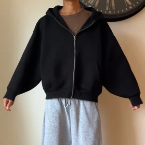 Svart oversized hoodie med dragkedja - Svart oversized hoodie med huva och hel dragkedja framtill. Tröjan har stora fickor på magen och breda muddar vid ärmslut och nederkant. Perfekt för en chill och avslappnad stil.