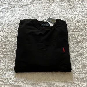 Svart sweatshirt från Polo Ralph Lauren - Svart sweatshirt från Polo Ralph Lauren med klassisk rund hals och lång ärm. Tröjan har det ikoniska röda broderade Polo-logot på bröstet och ribbade muddar vid ärmslut och nederkant. Perfekt för en clean och stilren look. Man kan gå  ner i pris!