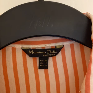 Randig skjorta från Massimo Dutti - Säljer en snygg skjorta från Massimo Dutti i vitt och orange röd färg med vertikala ränder. Skjortan har klassisk krage, långa ärmar och knappar framtill. Perfekt för dig som gillar stilrena och färgglada plagg.