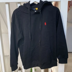 Ralph Lauren hoodie - Svart hoodie från Polo Ralph Lauren med röd broderad logga på bröstet. Tröjan har huva med snörning, känguruficka och långa ärmar. Tillverkad i mjuk bomull, perfekt för chill dagar. Klassisk och stilren design som funkar till allt.