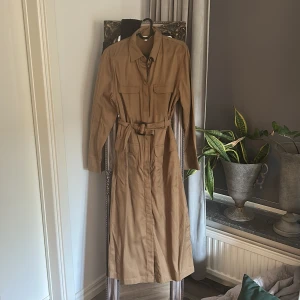 Beige långklänning med bälte från Indiska - Snygg beige långklänning från Indiska med långa ärmar, knappar framtill och bälte i midjan. Klänningen har stora fickor på bröstet och framtill, samt klassisk krage. Perfekt för dig som gillar stilrena och tidlösa plagg.
