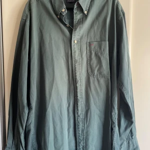 Gant bomullsskjorta grön stl M - Snygg grön skjorta från GANT i 100% bomull med klassisk button-down krage och bröstficka. Skjortan har ljusa knappar och diskreta röda detaljer vid fickan och ryggen. Perfekt för dig som gillar stilrena och tidlösa plagg. Välanvänd skön. Kamerans röda sökare orsakar det lite bleka uttrycket i mitten men skjortan är jämnt grön.