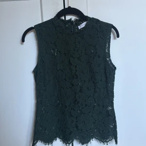 Mörkgrön spetsblus från IVY & OAK - Supersnygg mörkgrön ärmlös blus från IVY & OAK i spets med blommigt mönster. Toppen har hög hals, dragkedja bak och fina råa kanter nertill. Perfekt för dig som vill ha en elegant och unik look med extra detaljer.