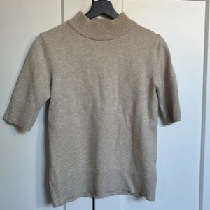 Beige polotröja från VILA - Snygg beige polotröja från VILA i stickad kvalitet. Tröjan har ribbade detaljer vid krage, ärmslut och nederkant samt halvlånga ärmar. Perfekt för dig som gillar en clean och enkel look med lite extra cozy vibe.