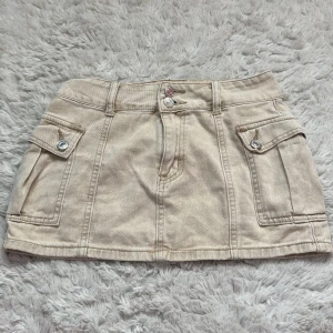 Beige jeanskjol från SHEIN XS - Superfin beige kort jeanskjol från SHEIN i storlek XS. Kjolen har två stora fickor med lock och silverfärgade knappar framtill, samt dubbla knappar i midjan. Snygg och enkel att matcha med olika toppar. Använt fåtal gånger och nypris är 179kr.
