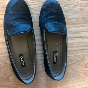 Svarta loafers i mocka från ECCO - Stilrena svarta loafers från ECCO i mjuk mocka med lätt glansig finish. Skorna har en klassisk rund tå och platt sula, perfekt för en clean och enkel look. Insidan är fodrad i beige och sulan är svart. Snygga till både jeans och dressade byxor.