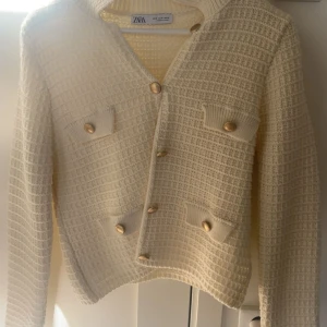Beige kofta från Zara med guldknappar - Snygg beige kofta från Zara med strukturerat stickat material och dekorativa guldknappar framtill. Koftan har fyra fickor med lock och långa ärmar. Perfekt för lager-på-lager och ger en chic vibe till din outfit.