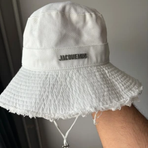 Vit Jacquemus bucket hat med snörning - Vit bucket hat från Jacquemus i bomull med bred brätte och fransad kant. Hatten har justerbar snörning med metall-detalj och Jacquemus-logga i silver framtill. Perfekt accessoar för en clean och trendig look.