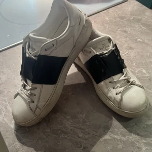 Valentino Garavani Open sneakers - Snygga Valentino Garavani Open sneakers i vitt skinn med bred svart läderdetalj över mitten. Klassisk rund tå, vita snören och platt sula. Kommer med original röd box och dustbag. Perfekta för dig som vill ha en lyxig och stilren look. Pris går att diskuteras 