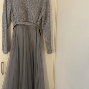 Lång grå klänning med glitterdetaljer - Superfin långklänning i grått med glittrig överdel och lång, flowy kjol i tunt tyg. Klänningen har långa ärmar och ett matchande bälte i midjan. Perfekt för dig som vill sticka ut med en elegant och stilren look.