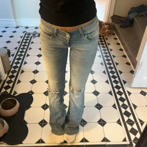 Ljusblå bootcut jeans med slitningar - Säljer ett par ljusblå jeans med bootcut-modell och slitna detaljer vid knäna. Jeansen har låg midja. Materialet är denim och passformen är normal med lätt utsvängda ben. 