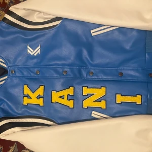 Karl Kani blå och vit bomberjacka - Blå och vit bomberjacka från Karl Kani med stora gula bokstäver framtill och en stor gul smiley på ryggen. Jackan har vita ärmar, blå kropp, ribbade muddar med ränder och tryckknappar. Materialet är syntet med skinnkänsla och detaljer i svart och vitt.