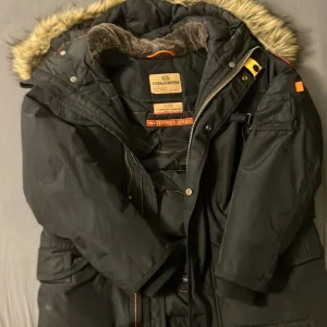 Svart Parajumpers parkas med fuskpäls - Grym svart parkas från Parajumpers med ikonisk patch på ärmen och fuskpäls runt huvan. Jackan har flera fickor, dragkedja och knappar framtill samt mjukt foder i kragen. Perfekt för kalla dagar och riktigt snygg streetstil.