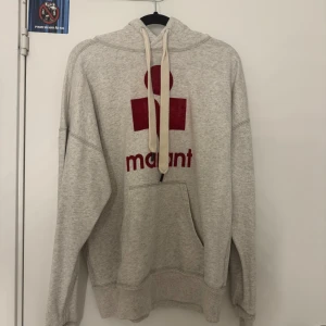 Grå hoodie från Isabel Marant - Grå hoodie från Isabel Marant med röd logga på bröstet. Tröjan har huva med snörning, stor magficka och långa ärmar. Mjuk bomullskvalitet och avslappnad passform, perfekt för chill dagar, men där du även vill se lyxig ut. 