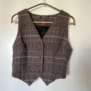 Rutig brun väst med knappar - Snygg rutig väst i bruna och beige toner med v-ringad skärning och knappar framtill. Västen har en skräddad passform och är fodrad på insidan. Perfekt för att ge outfiten en vintage och preppy vibe. Köpt second hand, saknar lapp men skulle säga att den passar xs/s. 