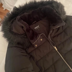 Svart pufferjacka med fuskpäls från ONLY - Svart pufferjacka från ONLY med mysig fuskpäls runt huvan och dragkedja framtill. Jackan har quiltad design, två fickor med tryckknappar och snygga detaljer i guld. Perfekt för kalla dagar och har en klassisk, lite kortare passform.