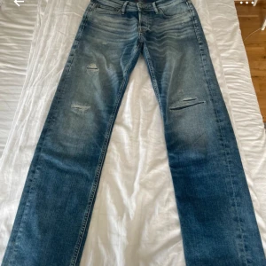 Blåa jeans från Jack & Jones - Snygga blå jeans från Jack & Jones i modellen Comfort/Mike. Jeansen har en rak passform med slitna detaljer och ljus tvätt. Klassisk femficksdesign, bälteshällor och dragkedjegylf. Perfekta för en avslappnad och trendig look.