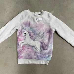 Pastellfärgad tröja med enhörning H&M - Supermjuk långärmad tröja från H&M i vitt med pastellfärgat motiv av en enhörning med glittriga paljettvingar. Tröjan har ribbade muddar och en fluffig känsla, perfekt för dig som älskar magiska vibes och färg.