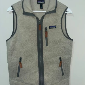 Beige fleeceväst från Patagonia XS - Mysig beige fleeceväst från Patagonia med grå detaljer och dragkedjor framtill. Västen har tre fickor med dragkedja och klassisk logga på bröstet. Perfekt för lager-på-lager och outdoor-vibe.