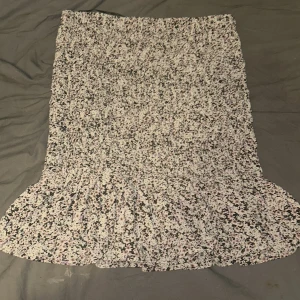 Blommig volangkjol från Vero Moda - Supersöt blommig kjol från Vero Moda i ljusa toner av beige, svart, lila och rosa. Kjolen har smockad midja och volang nertill för en extra flowy känsla. Perfekt för dig som gillar romantiska och trendiga plagg. Materialet är mjuk polyester.