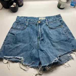 Blå jeansshorts med fransig kant - Säljer ett par ljusblå jeansshorts i klassisk modell med hög midja och rå, fransig kant. Shortsen har fem fickor och slitna detaljer, bland annat på bakfickan. Perfekta för sommaren och ger en avslappnad vibe.