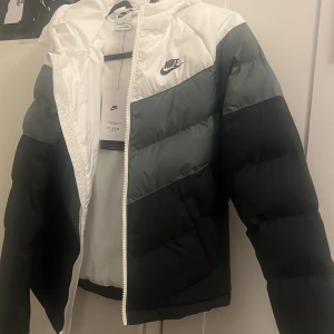 Nike pufferjacka svart/vit/grå L - Nike pufferjacka i färgblockad design med svart, grå och vit. Jackan har huva, dragkedja framtill och är vadderad med syntetfyllning. Klassisk Nike-logga på bröstet och sportig look. Perfekt för kalla dagar när du vill ha både stil och värme.