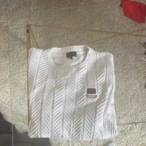 Vit stickad tröja från Morris - Säljer en vit stickad tröja från Morris med snyggt kabelstickat mönster och broderad patch med brittisk flagga på bröstet. Tröjan har rund halsringning, ribbade muddar och långa ärmar. Perfekt för dig som gillar klassisk och stilren look.