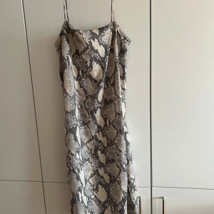 Beige och svart slipklänning med ormmönster - Säljer en snygg slipklänning med tunna axelband och trendigt ormmönster i beige och svart. Klänningen har en rak passform och faller snyggt längs kroppen. Perfekt för dig som vill sticka ut med ett djurmönster och en stilren look.