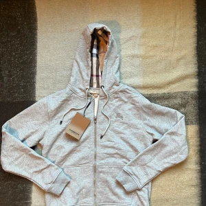 Grå hoodie från Burberry med dragkedja - Snygg grå hoodie från Burberry med klassiskt rutigt foder i huvan och broderad logga på bröstet. Tröjan har dragkedja framtill, snörning i huvan och två fickor. Perfekt för dig som vill ha en stilren och exklusiv look.