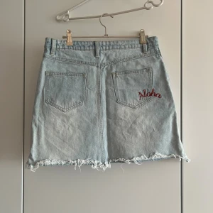 Broderad jeanskjol med fransig kant - Ljusblå jeanskjol med broderade blommor framtill och röd 'Aloha'-text på bakfickan. Kjolen har fransig, rå kant nertill och klassiska fickor både fram och bak. Perfekt för dig som vill ha en chill och trendig look med lite extra detaljer.