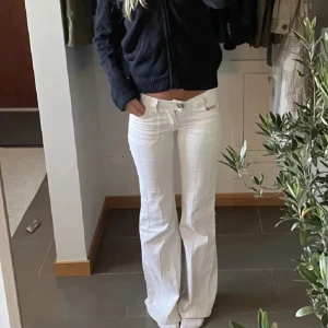 Vita bootcut byxor med rutigt mönster - Säljer ett par vita bootcut byxor med subtilt rutigt mönster. De har låg midja, framfickor och markerade sömmar som ger en snygg retrovibe. Materialet känns som bomull med lite stretch, perfekta för dig som gillar en avslappnad men trendig look.