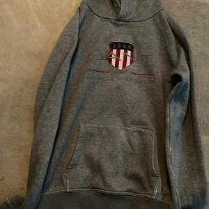 Grå hoodie från Gant med tryck - Säljer en grå hoodie från Gant med broderat emblem och text på bröstet. Tröjan har huva, magficka och långa ärmar. Perfekt för en avslappnad och sportig stil.
