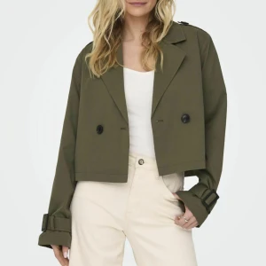 Olivgrön croppad trenchcoat - Snygg croppad trenchcoat i olivgrönt med bred krage, dubbelknäppning och coola spänndetaljer vid ärmsluten. Jackan har en loose fit och långa ärmar, perfekt för dig som vill ha en trendig och stilren look.