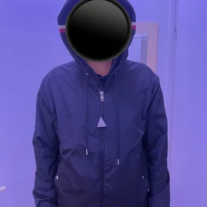 Moncler vindjacka med huva, svart - Svart vindjacka från Moncler med huva och dragkedja framtill. Jackan har Moncler-logga på ärmen och snygga röd-vita detaljer på huvan. Tillverkad i lätt polyester, perfekt för blåsiga dagar. Fickor med tryckknappar och justerbar dragsko vid huvan.