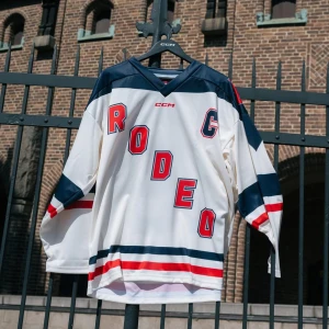 Vit och marinblå hockeytröja CCM - Säljer en vit och marinblå hockeytröja från CCM med röda och blå detaljer. Tröjan har tryck med texten 'RODEO' och 'BOLAGET' samt siffran 19 på ryggen och axlarna. Långärmad modell med breda ränder och sportig look. Perfekt för dig som gillar statement-plagg.