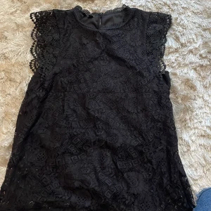 Svart spetsblus från Pieces XS - Supersnygg svart blus från Pieces i storlek XS. Blusen har vackra spetsdetaljer över hela plagget och fina virkade kanter vid ärmar och hals. Ärmlös modell med rund halsringning och knapp i nacken. Perfekt för dig som gillar detaljer och en elegant look.