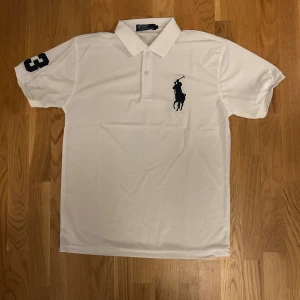 Vit pikétröja från Polo Ralph Lauren - Vit pikétröja från Polo Ralph Lauren med klassisk krage, knäppning framtill och stor marinblå logga på bröstet. Kortärmad modell med siffra 3 på ärmen. Perfekt för en clean och sportig stil.