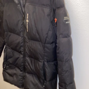 Svart pufferjacka från Sail Racing - Svart pufferjacka från Sail Racing med markerad logga på bröstet och ärmen. Jackan har dragkedja framtill, två sidofickor med dragkedja och lång ärm. Perfekt för kalla dagar med sin vadderade design och sportiga look.