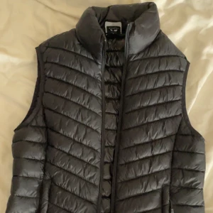 Svart dunväst från McKinley, strl 36 - Snygg svart dunväst från McKinley i storlek 36. Västen har quiltad design, hög krage och dragkedja framtill. Perfekt för lager-på-lager och outdoor-vibes. Materialet är glansigt och lätt, vilket gör den smidig att bära och packa ner.