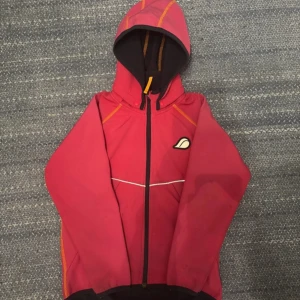 Röd vindjacka från Didriksons, strl 130 - Röd vindjacka från Didriksons med huva, svarta och orange detaljer samt reflexband över bröstet. Jackan har hel dragkedja framtill och är tillverkad i 100% polyester. Perfekt för blåsiga dagar och har en sportig vibe.