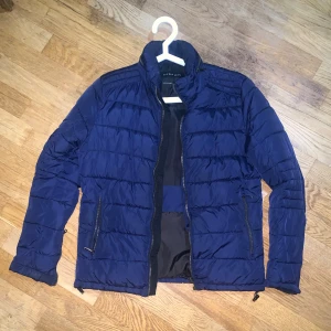 Blå pufferjacka från Zara Man - Snygg blå pufferjacka från Zara Man med horisontella sömmar, hög krage och dragkedjor både framtill och på fickorna. Jackan är lätt vadderad och har en stilren look som passar perfekt till kyligare dagar.